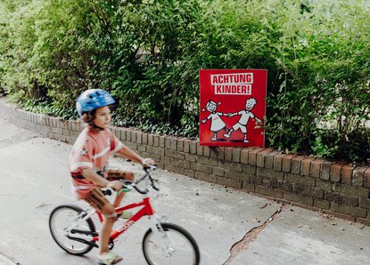 Achtung Kinder Plakat aus dem Jahr 2021 im Vordergrund fährt Kind auf Fahrrad vorbei
