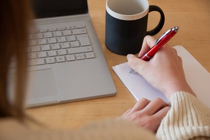 Jemand schreibt mit einem roten Kugelschreiber auf einen Block auf einem Schreibtisch.