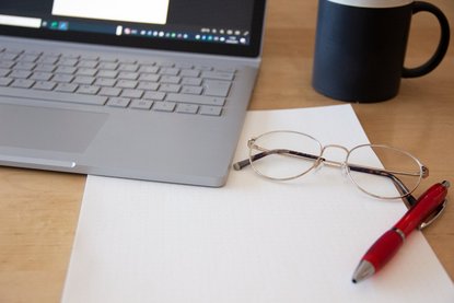 Brille, Stift, Block, Laptop und Kaffeetasse auf dem Schreibtisch.